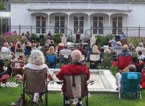 A past Poetry in the Garden event at the Keeler Tavern Museum. Guests sitting in lawn chairs with poet at microphone sharing their work.